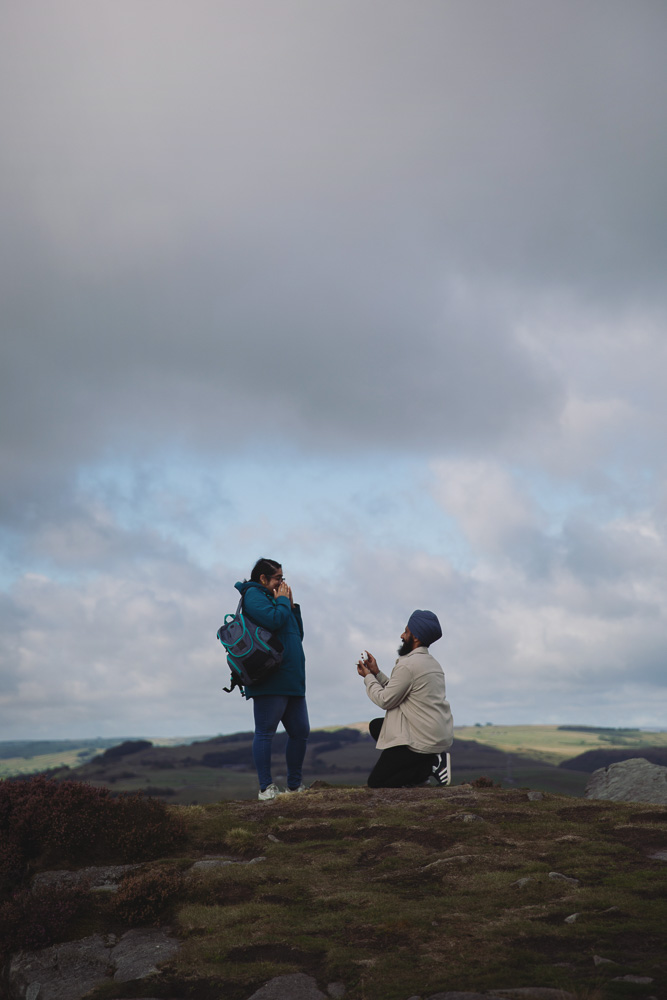 Surprise proposal in the Peak District as a man kneels to propose to his partner with the countryside in the background